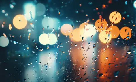 Camera pans across a rain droplet covered window with city lights illuminating behind