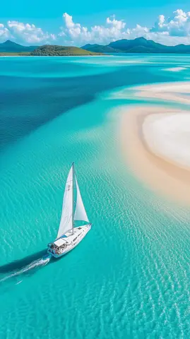 A sailboat glides across tranquil turquoise waters in a pristine tropical lagoon surrounded by lush green hills and fluffy white clouds.